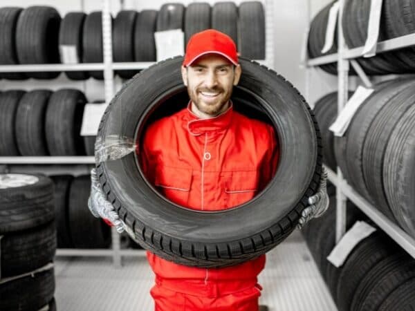 Un tecnico di un deposito gomme, in tuta rossa e cappellino, sorride mentre tiene un pneumatico, con scaffali pieni di gomme in background.